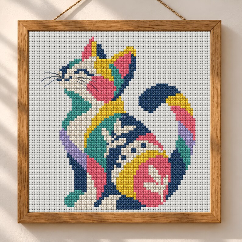 May include: A square cross-stitch embroidery of a cat in a wooden frame. The cat is composed of geometric color blocks in navy blue, bright yellow, pink, teal, and lavender, featuring white leaf-like patterns on its body and a white background.