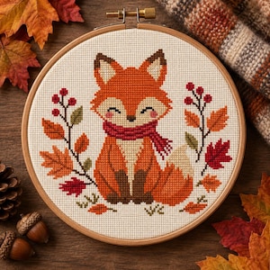 May include: A circular wooden embroidery hoop containing a cross-stitch pattern of a fox with a red scarf, surrounded by autumn leaves and red berries on a cream-colored fabric background. The fox has orange fur, brown paws, and a white tail tip.