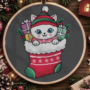 May include: A cross-stitch embroidery in a wooden hoop featuring a white cat with blue eyes wearing a red and green elf hat. The cat sits inside a red Christmas stocking with white trim, surrounded by wrapped gifts and green holly leaves.