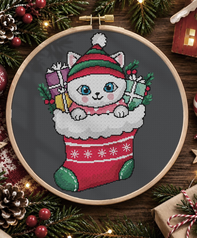 May include: A cross-stitch embroidery in a wooden hoop featuring a white cat with blue eyes wearing a red and green elf hat. The cat sits inside a red Christmas stocking with white trim, surrounded by wrapped gifts and green holly leaves.