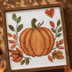 Puede incluir: Un bordado de punto de cruz cuadrado que presenta una calabaza naranja central con un tallo verde, rodeada de hojas verdes y follaje otoñal naranja. Un pequeño corazón naranja se sitúa sobre la calabaza, todo en un marco de madera.