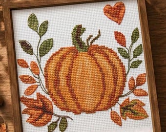 May include: A square cross-stitch embroidery featuring a central orange pumpkin with a green stem, surrounded by green leaves and orange autumn foliage. A small orange heart is positioned above the pumpkin, all encased in a wooden frame.