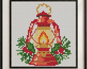 May include: A framed cross-stitch embroidery featuring a red lantern with a glowing yellow and orange flame at its center. The lantern is flanked by green holly leaves and small red berries on both sides, all set against a white grid fabric.
