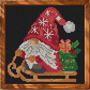 May include: A square cross-stitch art piece in a wooden frame featuring a gnome wearing a red hat with white snowflake patterns, a white beard, and a tan nose. The gnome sits on a tan wooden sled next to a green gift bag with red berries.