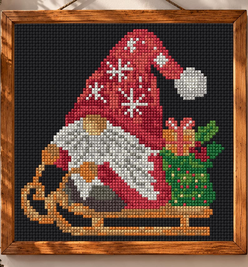 May include: A square cross-stitch art piece in a wooden frame featuring a gnome wearing a red hat with white snowflake patterns, a white beard, and a tan nose. The gnome sits on a tan wooden sled next to a green gift bag with red berries.