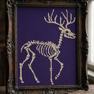 May include: A cross-stitch embroidery featuring a beige deer skeleton on a dark purple fabric background, encased in an ornate, dark brown wooden frame with intricate carved details along the edges and corners of the rectangular border.