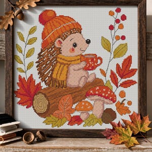 May include: A cross-stitch embroidery in a dark wood frame depicting a hedgehog wearing an orange knit hat and yellow scarf, holding a red mug. The scene includes a brown log, two spotted mushrooms, autumn leaves, and red berries on a white grid.