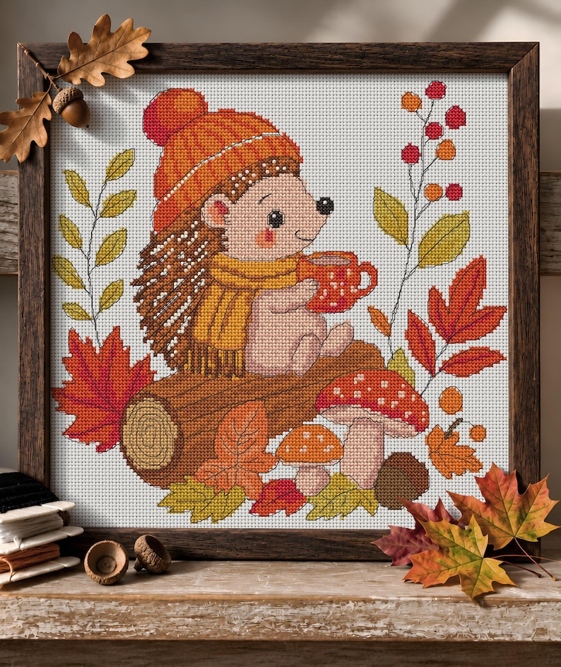 May include: A cross-stitch embroidery in a dark wood frame depicting a hedgehog wearing an orange knit hat and yellow scarf, holding a red mug. The scene includes a brown log, two spotted mushrooms, autumn leaves, and red berries on a white grid.