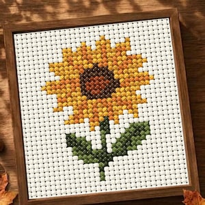 May include: A square cross-stitch embroidery featuring a single sunflower with a dark brown center and bright yellow petals, set against a white Aida cloth background. The piece is enclosed in a simple, thin, medium-toned wooden frame.