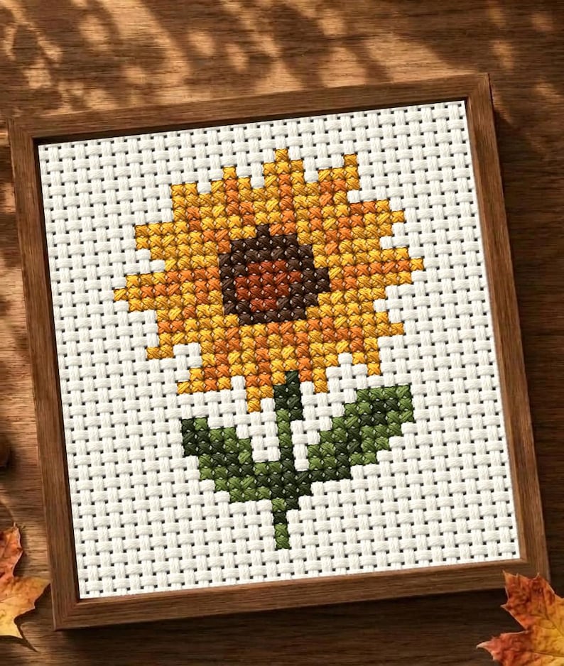 May include: A square cross-stitch embroidery featuring a single sunflower with a dark brown center and bright yellow petals, set against a white Aida cloth background. The piece is enclosed in a simple, thin, medium-toned wooden frame.