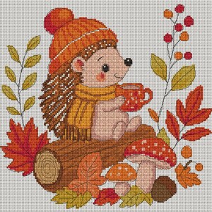 May include: A cross-stitch pattern featuring a hedgehog wearing an orange knit hat and yellow scarf, holding a red mug. The hedgehog sits on a brown log surrounded by red and yellow autumn leaves, two red-capped mushrooms, and a berry sprig.