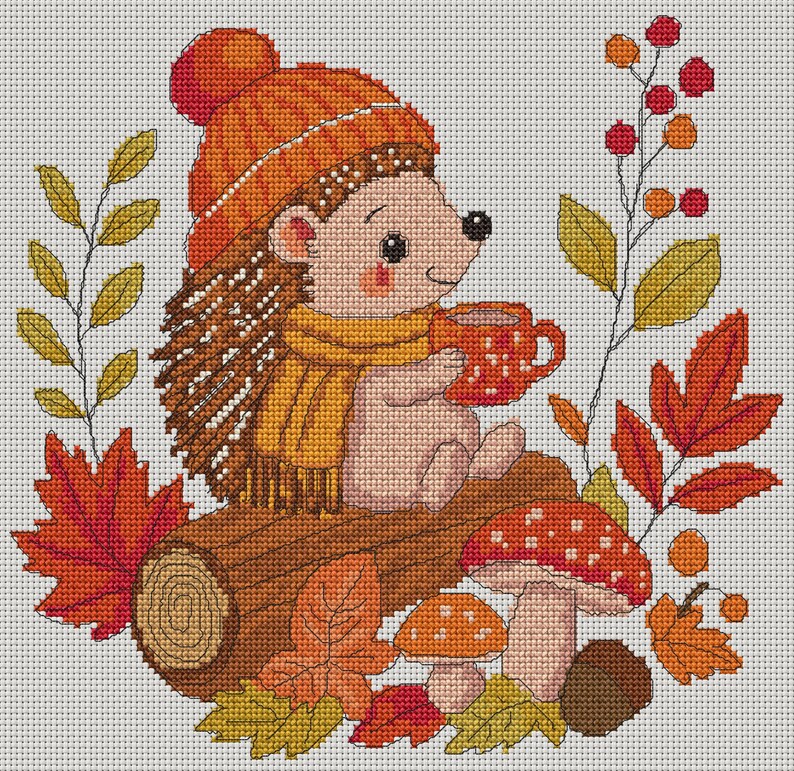 May include: A cross-stitch pattern featuring a hedgehog wearing an orange knit hat and yellow scarf, holding a red mug. The hedgehog sits on a brown log surrounded by red and yellow autumn leaves, two red-capped mushrooms, and a berry sprig.