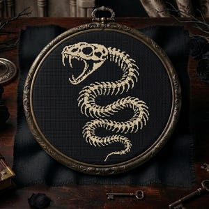 May include: A circular embroidery hoop containing a black fabric piece with a cross-stitched design of a beige snake skeleton. The snake is depicted in a coiled position with its mouth open, showing fangs. The hoop has an ornate, dark metal frame.