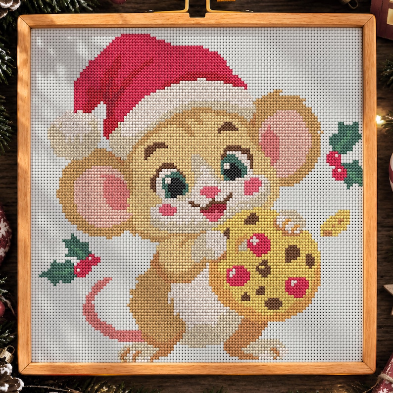 Op de afbeelding: Een kruissteekborduurwerk in een houten lijst met een beige muis die een rode kerstmuts draagt. De muis heeft grote oren, groene ogen, roze wangen en houdt een geel koekje met chocoladestukjes vast. Groene hulstblaadjes met rode bessen zijn zichtbaar.