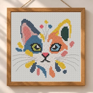 K&ouml;nnte beinhalten: Ein quadratisches Kreuzstich-Stickbild eines Katzengesichts in einem Holzrahmen. Die Katze hat eine wei&szlig;e Grundfarbe mit blauen, orangefarbenen, gelben und rosafarbenen Flecken. Sie hat ein gr&uuml;nes und ein blaues Auge, eine rosa Nase und schwarze Schnurrhaare.