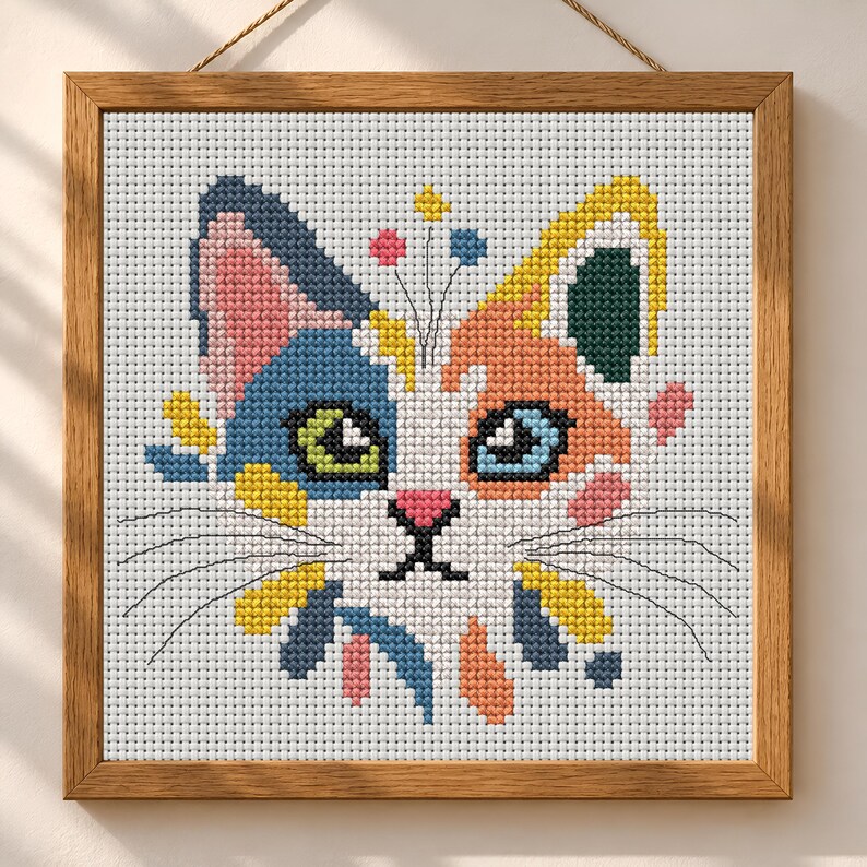 K&ouml;nnte beinhalten: Ein quadratisches Kreuzstich-Stickbild eines Katzengesichts in einem Holzrahmen. Die Katze hat eine wei&szlig;e Grundfarbe mit blauen, orangefarbenen, gelben und rosafarbenen Flecken. Sie hat ein gr&uuml;nes und ein blaues Auge, eine rosa Nase und schwarze Schnurrhaare.