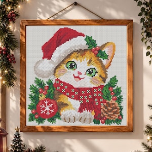 May include: A square cross-stitch embroidery in a wooden frame depicting a kitten wearing a red and white Santa hat and a red scarf with white dots. The kitten is surrounded by green holly leaves, red berries, a red ornament with a white snowflake, and a brown pinecone.