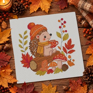 May include: A cross-stitch pattern on white fabric featuring a hedgehog wearing an orange knit hat and yellow scarf, holding a red mug. The design includes a brown log, red and orange autumn leaves, two mushrooms, and a sprig of red berries.