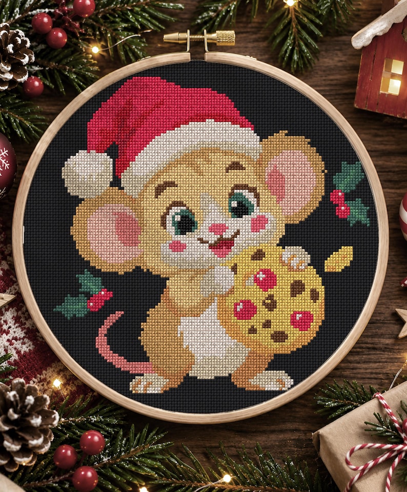 Op de afbeelding: Een kruissteekborduurwerk in een ronde houten ring met een beige muis met grote oren en roze wangen die een rode kerstmuts draagt. De muis houdt een geel koekje met rode en bruine stippen vast tegen een zwarte achtergrond met hulsttakjes.