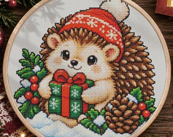 May include: A cross-stitch embroidery in a round wooden hoop featuring a hedgehog wearing a red winter hat with a white pom-pom. The hedgehog holds a green gift box with a red ribbon, surrounded by holly leaves, red berries, and a pinecone.