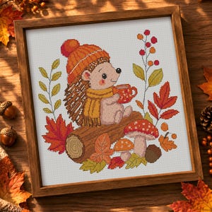 May include: A framed cross-stitch embroidery featuring a hedgehog wearing an orange knit hat and yellow scarf, holding a red mug. The design includes a brown log, red and orange autumn leaves, two mushrooms, and a sprig of red berries on white fabric.