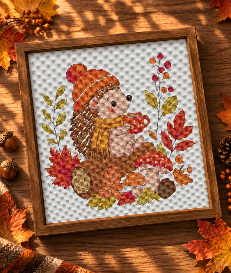 May include: A framed cross-stitch embroidery featuring a hedgehog wearing an orange knit hat and yellow scarf, holding a red mug. The design includes a brown log, red and orange autumn leaves, two mushrooms, and a sprig of red berries on white fabric.