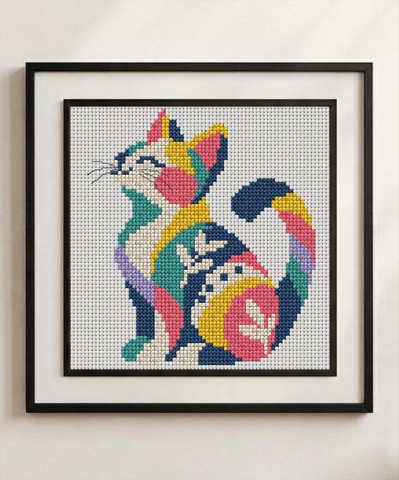 May include: A framed cross-stitch embroidery featuring a stylized cat composed of geometric color blocks in shades of navy blue, bright pink, yellow, teal, and lavender against a white grid background, enclosed in a simple black frame.