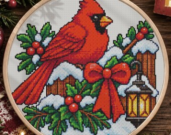 May include: A circular wooden embroidery hoop containing a cross-stitch pattern of a bright red cardinal bird perched on a snow-covered wooden fence. The design includes green holly leaves, red berries, a large red bow, and a lit lantern.