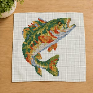 May include: A cross-stitch embroidery piece on white Aida fabric featuring a colorful fish leaping upward. The fish is rendered in shades of green, orange, yellow, and blue, with a detailed eye and open mouth, showcasing intricate needlework stitches.