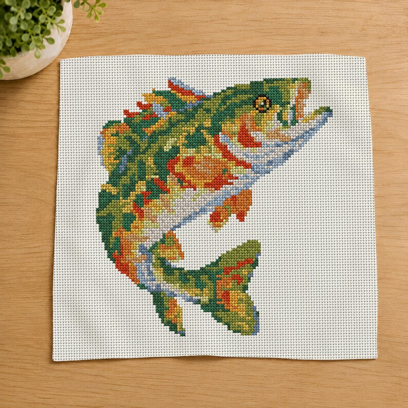 May include: A cross-stitch embroidery piece on white Aida fabric featuring a colorful fish leaping upward. The fish is rendered in shades of green, orange, yellow, and blue, with a detailed eye and open mouth, showcasing intricate needlework stitches.
