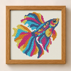 May include: A framed cross-stitch embroidery featuring a vibrant betta fish with flowing fins in shades of magenta, bright blue, yellow, and orange. The fish is depicted against a white grid fabric background, enclosed in a light brown wooden frame.