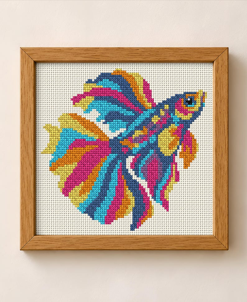 May include: A framed cross-stitch embroidery featuring a vibrant betta fish with flowing fins in shades of magenta, bright blue, yellow, and orange. The fish is depicted against a white grid fabric background, enclosed in a light brown wooden frame.