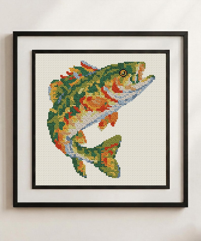May include: A framed cross-stitch embroidery featuring a colorful fish in shades of green, orange, yellow, and blue. The fish is depicted in a leaping position against a cream-colored fabric background, enclosed within a simple black frame.
