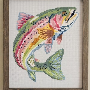 May include: A cross-stitch embroidery of a rainbow trout featuring vibrant green, pink, and yellow thread colours on white fabric. The fish is depicted in a leaping motion and is enclosed within a simple, dark brown wooden frame with a square shape.
