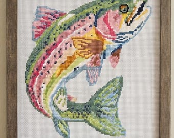 May include: A cross-stitch embroidery of a rainbow trout featuring vibrant green, pink, and yellow thread colours on white fabric. The fish is depicted in a leaping motion and is enclosed within a simple, dark brown wooden frame with a square shape.