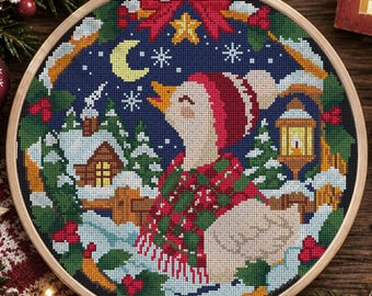 May include: A circular wooden embroidery hoop containing a cross-stitch pattern of a white goose wearing a red and green plaid scarf and a red hat. The scene features a snowy winter landscape with a cabin, a yellow lantern, and a crescent moon.
