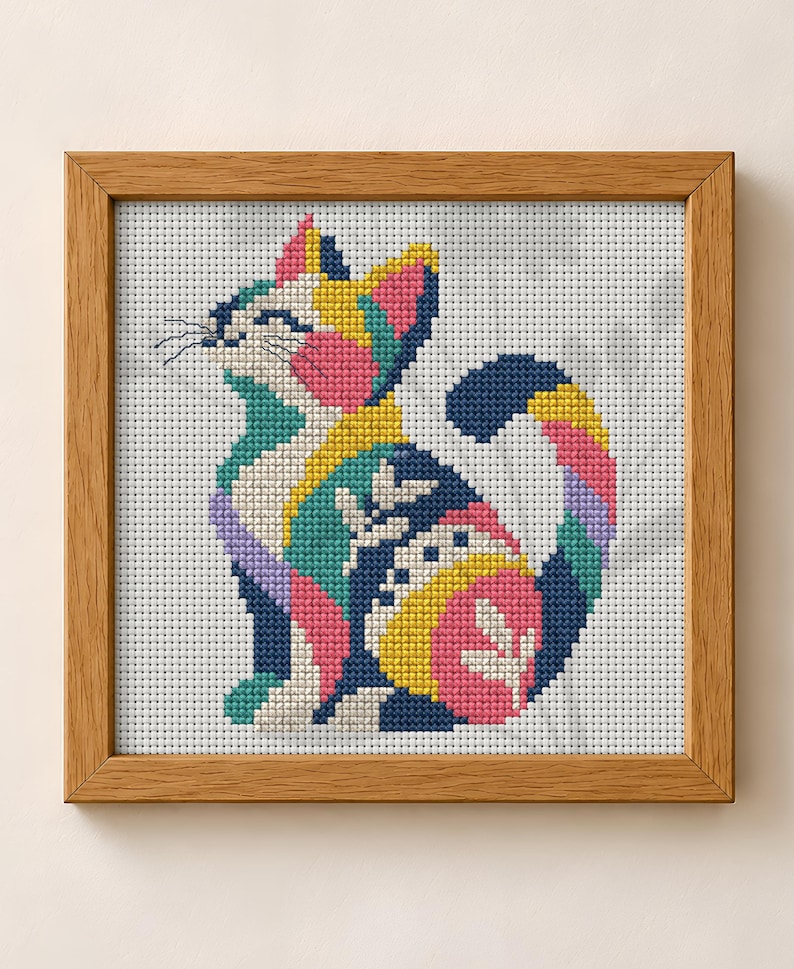 May include: A cross-stitch embroidery of a cat in a wooden frame. The cat is composed of geometric color blocks in navy blue, bright yellow, pink, teal, and white, featuring stylized leaf patterns on its body and a curved, multi-colored tail.