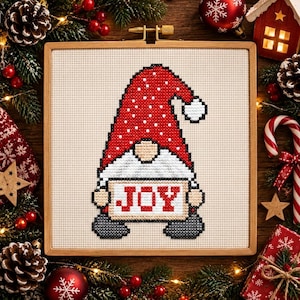 May include: A cross-stitch embroidery in a square wooden hoop featuring a gnome with a red hat covered in white dots, a white beard, and gray boots. The gnome holds a rectangular sign with the word JOY stitched in red letters on a white background.