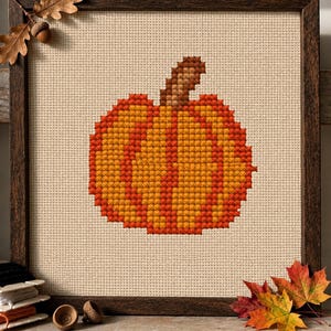 May include: A cross-stitch embroidery of an orange and yellow pumpkin with a brown stem, centered on a beige fabric background. The piece is enclosed in a dark brown wooden frame, accented by a small oak leaf and acorn in the top corner.