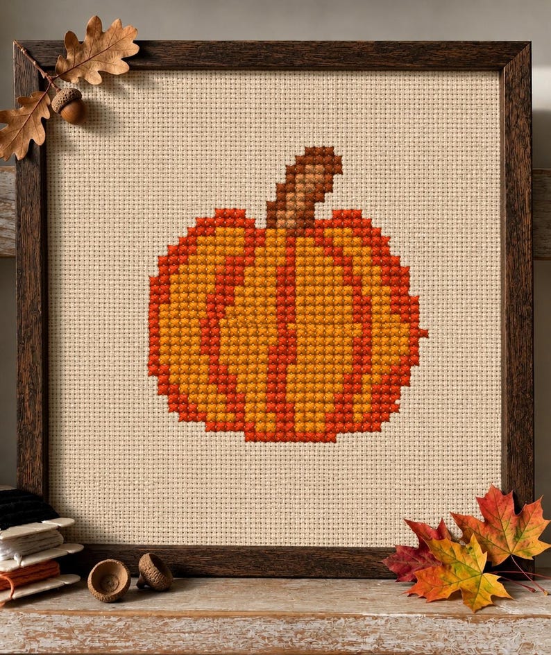 May include: A cross-stitch embroidery of an orange and yellow pumpkin with a brown stem, centered on a beige fabric background. The piece is enclosed in a dark brown wooden frame, accented by a small oak leaf and acorn in the top corner.