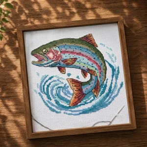 May include: A framed cross-stitch embroidery featuring a colourful rainbow trout leaping out of stylised blue water. The fish displays shades of green, pink, and orange, set against a white fabric background within a simple wooden square frame.