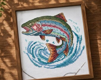 May include: A framed cross-stitch embroidery featuring a colorful rainbow trout leaping out of stylized blue water. The fish displays shades of green, pink, and orange, set against a white fabric background within a simple wooden square frame.