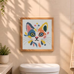 K&ouml;nnte beinhalten: Ein quadratisches Kreuzstich-Stickbild eines Katzengesichts in einem hellen Holzrahmen. Die Katze hat eine wei&szlig;e Basis mit blauen, orangefarbenen, gelben und rosafarbenen Flecken. Sie hat gr&uuml;ne und blaue Augen, eine rosa Nase und d&uuml;nne schwarze Schnurrhaare.