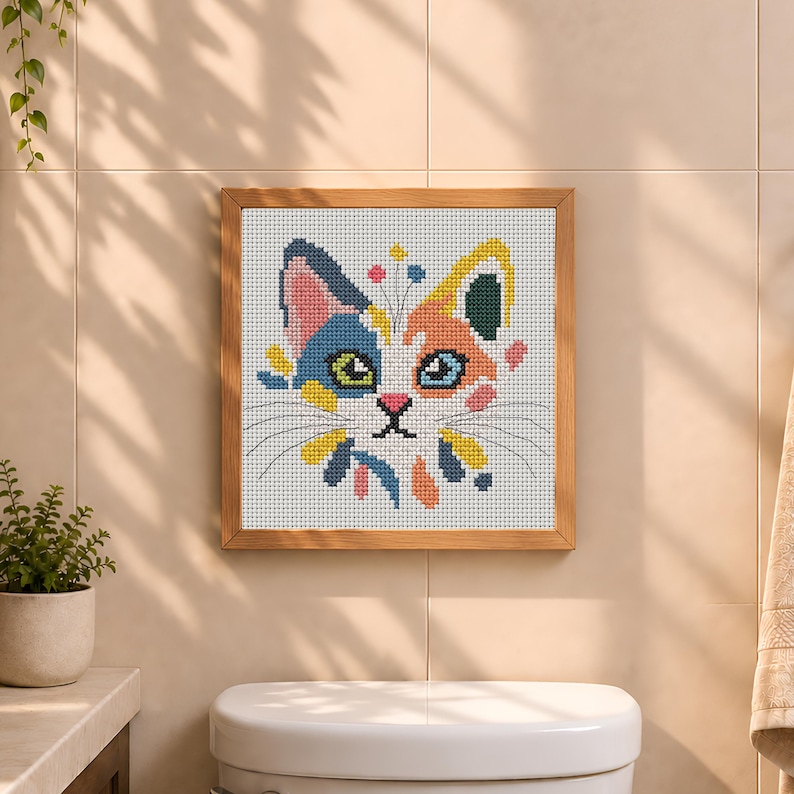 K&ouml;nnte beinhalten: Ein quadratisches Kreuzstich-Stickbild eines Katzengesichts in einem hellen Holzrahmen. Die Katze hat eine wei&szlig;e Basis mit blauen, orangefarbenen, gelben und rosafarbenen Flecken. Sie hat gr&uuml;ne und blaue Augen, eine rosa Nase und d&uuml;nne schwarze Schnurrhaare.
