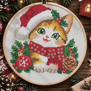 May include: A cross-stitch embroidery in a round wooden hoop featuring a kitten wearing a red and white Santa hat and a red scarf with white dots. The kitten is surrounded by green pine branches, red berries, a red ornament with a white snowflake, and a pinecone.