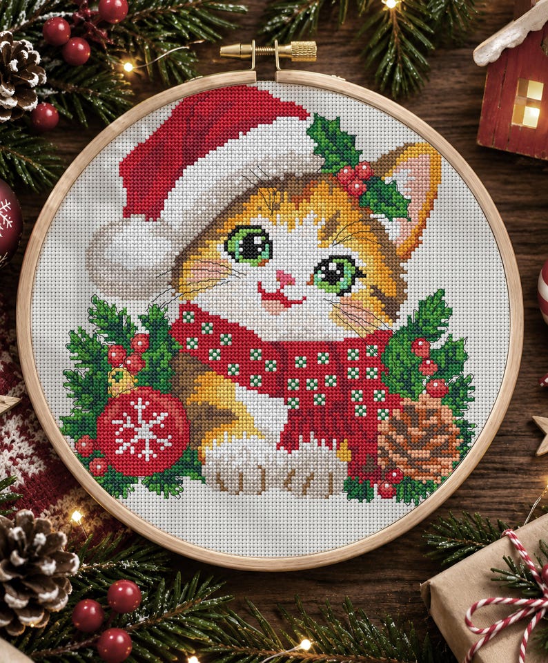 May include: A cross-stitch embroidery in a round wooden hoop featuring a kitten wearing a red and white Santa hat and a red scarf with white dots. The kitten is surrounded by green pine branches, red berries, a red ornament with a white snowflake, and a pinecone.