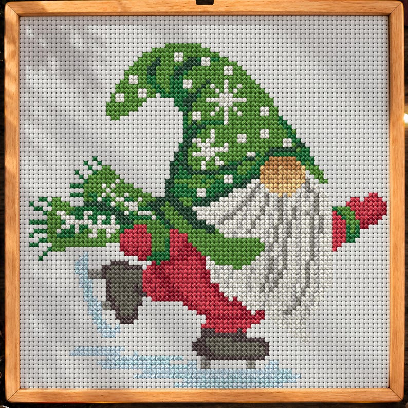 May include: A cross-stitch embroidery in a wooden frame depicting a gnome wearing a green hat with white snowflake patterns, a red outfit, and dark skates, gliding on ice while holding a red heart shape against a light gray background.