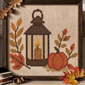 May include: A framed cross-stitch embroidery featuring a dark brown lantern with a yellow candle inside, flanked by an orange pumpkin and autumn leaves in shades of gold, brown, and orange, all set against a light beige fabric background.