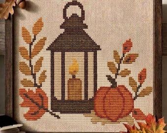 May include: A framed cross-stitch embroidery featuring a dark brown lantern with a yellow candle inside, flanked by an orange pumpkin and autumn leaves in shades of gold, brown, and orange, all set against a light beige fabric background.