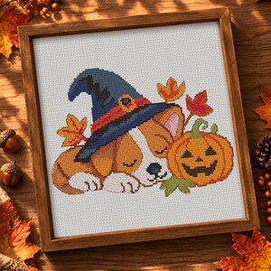 May include: A cross-stitch embroidery in a wooden frame featuring a sleeping puppy wearing a blue witch hat with an orange band. The puppy is positioned next to a carved orange jack-o'-lantern, surrounded by several small, colourful autumn leaves.